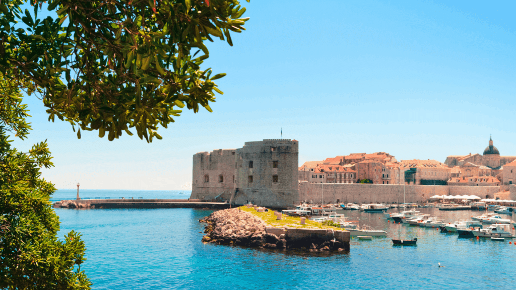 Sailboats and yachts in a turquoise Adriatic harbor beside Croatia’s historic stone fortifications and red-roofed cityscape.