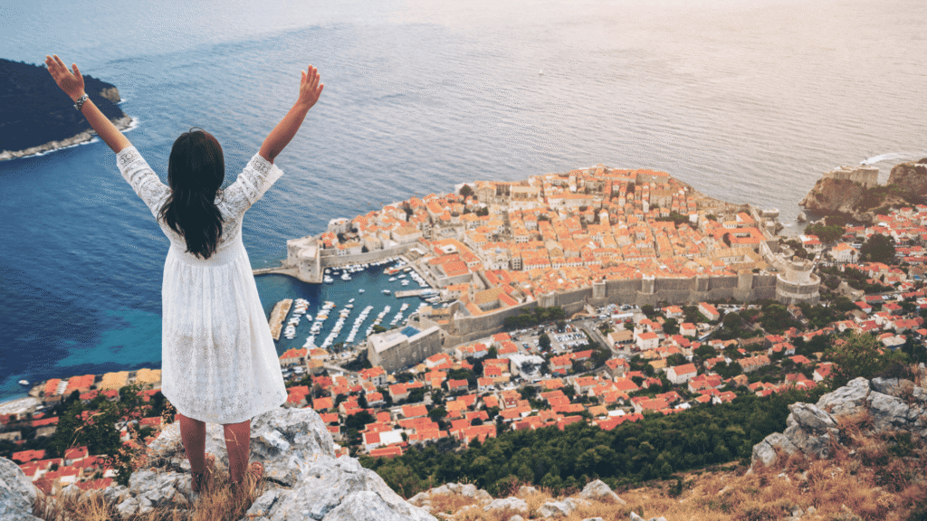 Traveler overlooking Dubrovnik’s historic city walls, red-roofed buildings and Adriatic coastline from a scenic hillside viewpoint.