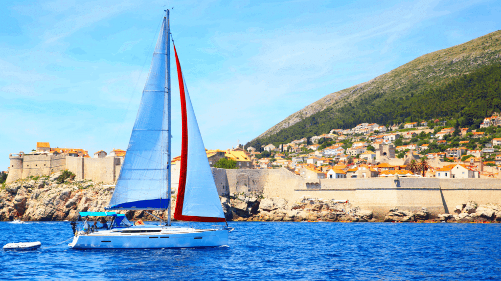 Sailboat cruising the blue Adriatic Sea beside Dubrovnik’s historic city walls and coastal landscape in Croatia.